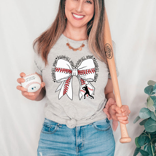 Woman wearing a gray t-shirt with a baseball and bow design, holding a baseball and a bat.