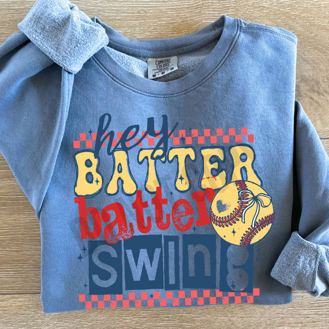 Blue sweatshirt with 'hey batter batter swings' text and softball graphic on a wooden surface