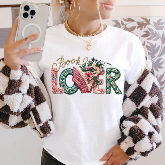 Person wearing a white sweatshirt with 'Book Lover' text and floral designs, holding a phone.