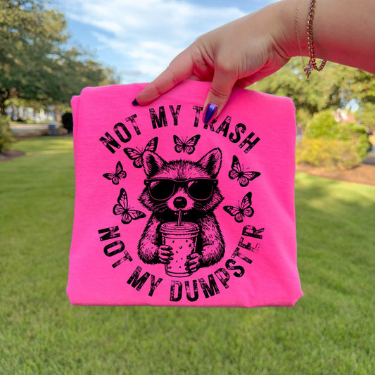Pink tote bag with a raccoon graphic and text held by a hand outdoors.