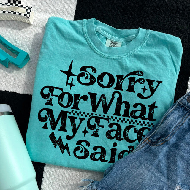 Turquoise t-shirt with black text 'Sorry For What My Face Said' on a checkered surface.
