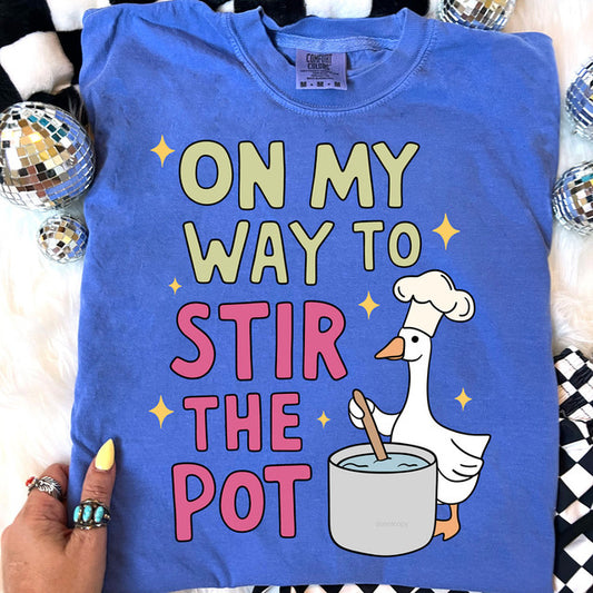 Blue t-shirt with a goose graphic and text 'On my way to stir the pot' on a decorative background.