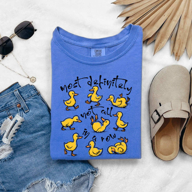 Blue t-shirt with yellow duck illustrations and text on a white surface with denim shorts, sunglasses, and clogs.