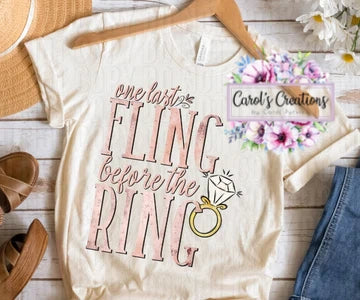 White t-shirt with 'one last fling before the ring' text and floral design on a wooden surface.
