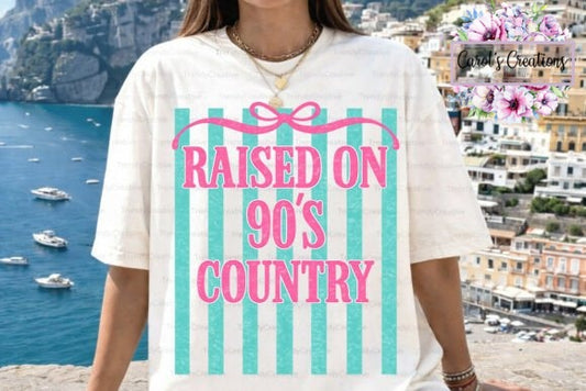 A woman wearing a white graphic t-shirt with a pink and teal design that reads 'RAISED ON 90'S COUNTRY'. The background shows a scenic coastal town with blue water and cliffs, and a 'Candy's Creations' watermark is visible in the top right corner.