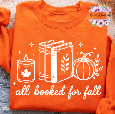 An orange crewneck sweatshirt featuring a white graphic print of a candle, books, a pumpkin, and the text 'all booked for fall'. The sweatshirt is laid flat on a textured beige surface with a denim jacket visible on the left side.