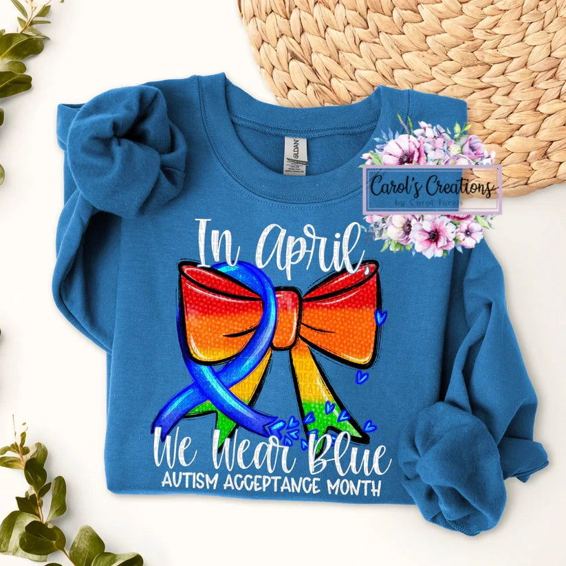 Blue sweatshirt with a colorful bow graphic and text about Autism Acceptance Month, displayed on a white background.