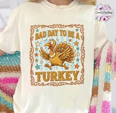 A cream-colored short-sleeve t-shirt featuring a cartoon turkey graphic and the text 'BAD DAY TO BE A TURKEY' in brown and gold, worn by a person holding a colorful striped knit blanket.