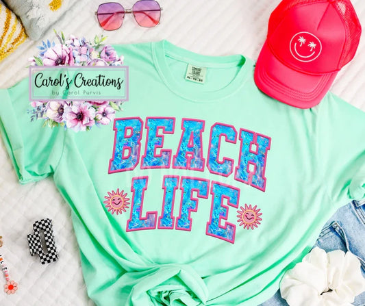 Mint green t-shirt with 'Beach Life' text, pink sunglasses, red cap, and denim shorts on a white surface.