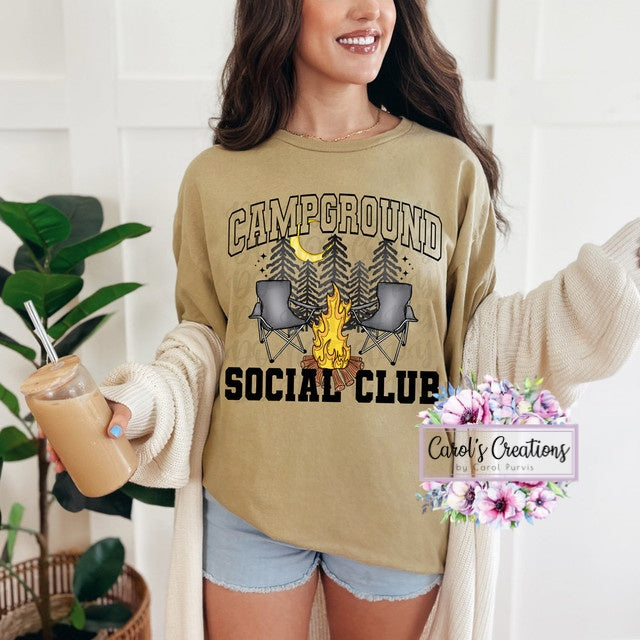 Woman wearing a 'Campground Social Club' shirt with a graphic of chairs around a campfire, holding a drink.