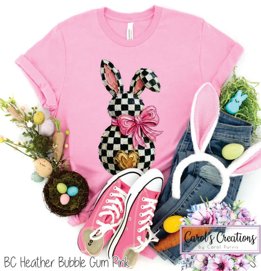 Pink t-shirt with checkered bunny design, pink sneakers, denim shorts, and Easter decorations on a white background.