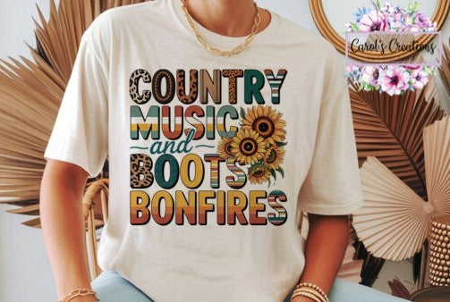 A person wearing a cream-colored graphic t-shirt with the text 'COUNTRY MUSIC and BOOTS BONFIRES' in a rustic multicolor font, decorated with sunflower graphics. The background features decorative dried palm leaves and a floral watermark logo for 'Carole's Creations'.