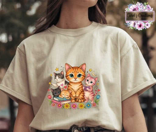 A person wearing a cream-colored t-shirt with a colorful cartoon graphic of an orange tabby cat and other cats sitting on books with flowers. A 'Carol's Creations' brand watermark is visible in the top right corner.