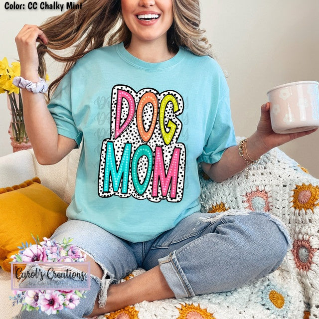 Woman wearing a 'Dog Mom' shirt sitting on a couch with a cup and flowers.