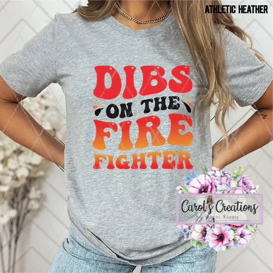 Gray t-shirt with colorful text 'DIBS ON THE FIRE FIGHTER' worn by a person, with a blurred background.