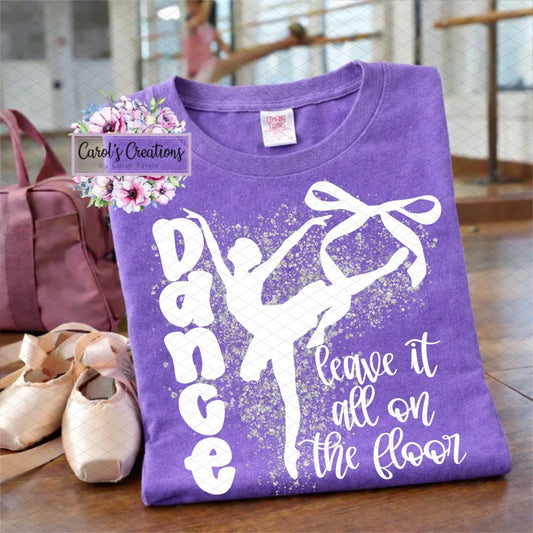 Purple t-shirt with a ballet design and text, placed on a wooden floor with ballet shoes and a pink bag.