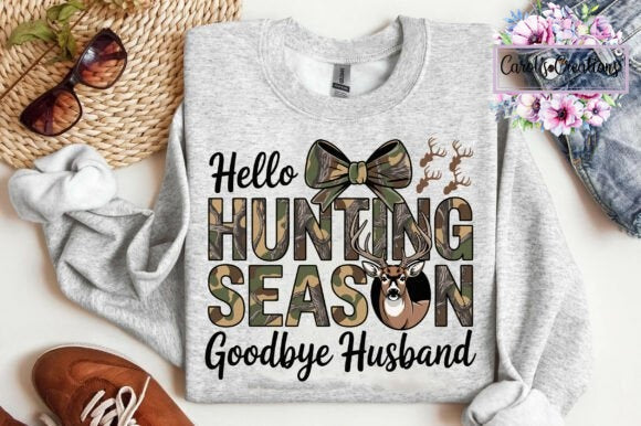 A heather gray long-sleeve crewneck sweatshirt laid flat with a hunting-themed graphic that reads 'Hello HUNTING SEASON Goodbye Husband' in black and camouflage lettering, plus a bow tie, deer head, and antler motifs. The sweatshirt is styled with sunglasses, a woven hat, a white towel, a brown shoe, and denim shorts on a light background.