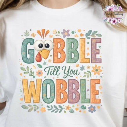 A white short-sleeve t-shirt featuring a colorful graphic that reads 'GOBBLE Till You WOBBLE' with a cartoon turkey face and floral accents, worn by a model.