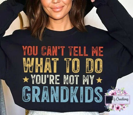 A woman wearing a black crewneck sweatshirt with a humorous graphic that reads 'YOU CAN'T TELL ME WHAT TO DO YOU'RE NOT MY GRANDKIDS' in red, yellow, and blue distressed lettering.