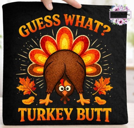A black square fabric item, likely a pillow cover or blanket, held by a hand at the top edge. The fabric features a graphic design with the text 'GUESS WHAT? TURKEY BUTT' in orange and yellow, surrounding a cartoon turkey with a brown body and orange and yellow tail feathers, accented with orange autumn leaves.