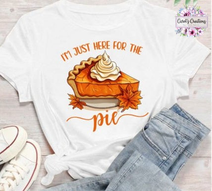 A white graphic t-shirt featuring an illustration of a slice of pumpkin pie with the text 'I'M JUST HERE FOR THE pie' in orange. The shirt is styled in a flat lay with light blue jeans, white sneakers, and a light wood background.