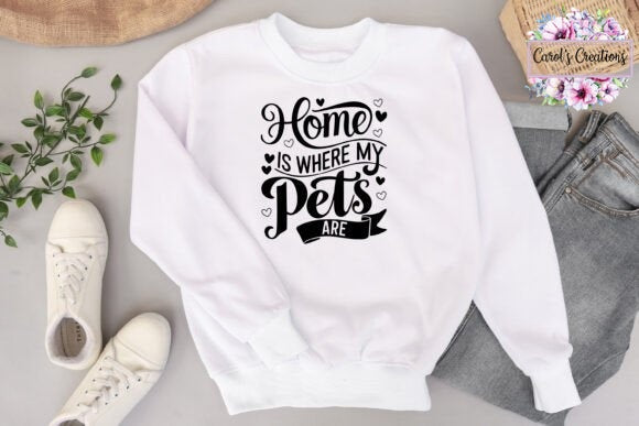 A white crewneck sweatshirt with a black graphic text reading 'Home IS WHERE MY Pets ARE' laid flat on a light gray surface, surrounded by white sneakers, gray jeans, a small green plant, a woven hat, and a floral card that says 'Carol's Christian's'.