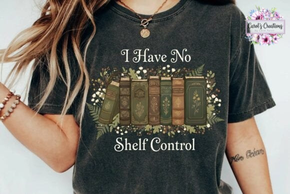 A person wearing a dark charcoal gray graphic t-shirt with the text 'I Have No Shelf Control' and an illustration of vintage books surrounded by flowers. The brand logo 'Carol's Creations' is visible in the top right corner.
