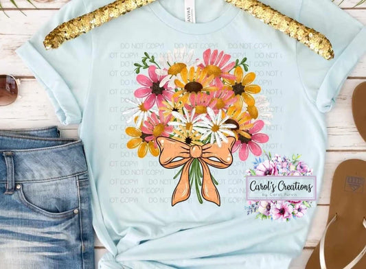 Light blue t-shirt with floral design and bow, placed on a wooden surface with jeans and sandals.