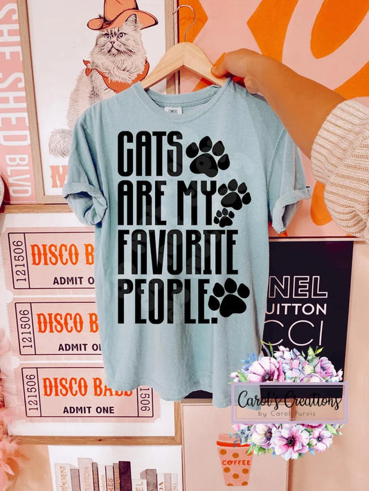 Light blue t-shirt with 'Cats are my favorite people' text and paw prints on a hanger against a colorful background.
