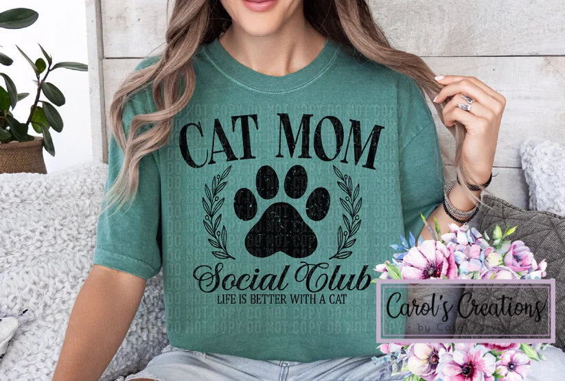 Person wearing a green t-shirt with 'Cat Mom Social Club' design, sitting on a couch with flowers and a plant in the background.