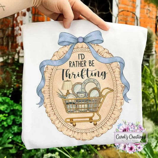 White pillow with 'I'd Rather Be Thrifting' design held by a hand outdoors.