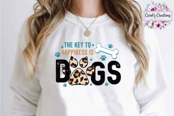A woman wearing a white long-sleeve sweatshirt with a graphic that reads 'THE KEY TO HAPPINESS IS DOGS' in black, blue, and tan leopard print. A 'Carol's Creations' logo is visible in the top right corner.