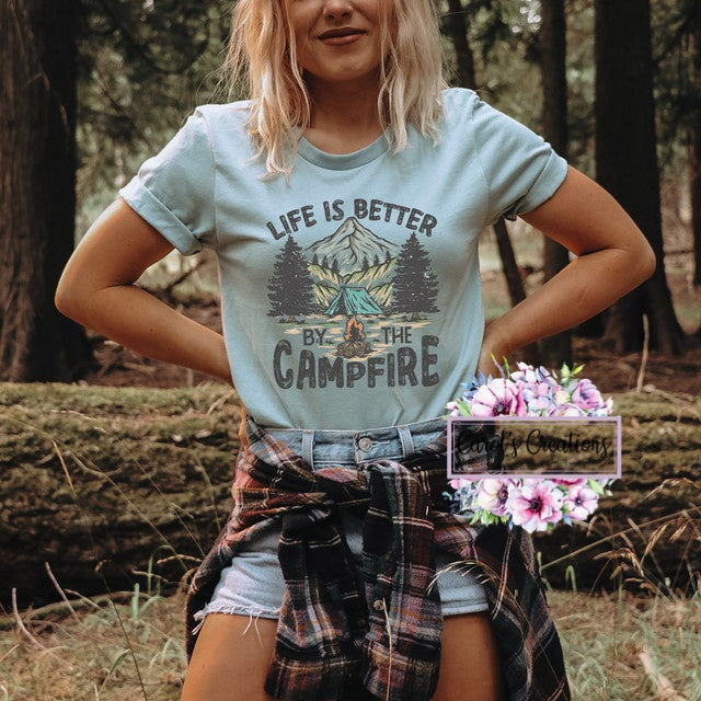 Person wearing a t-shirt with 'Life is Better by the Campfire' design in a forest setting.
