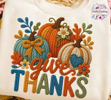 A close-up of a white garment featuring a colorful autumn-themed graphic with three pumpkins, leaves, flowers, and the words “give THANKS” in multicolored embroidered-style lettering. A small “Carly’s Creations” watermark appears in the top right corner.