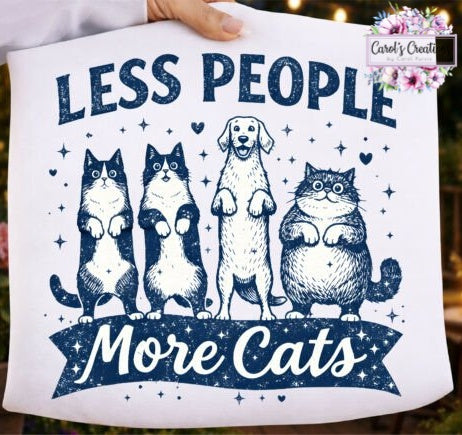 A white graphic t-shirt held up by a hand, featuring a navy blue design with the text 'LESS PEOPLE More Cats' and illustrations of four cats and a dog. A 'Carol's Creations' logo is visible in the top right corner.