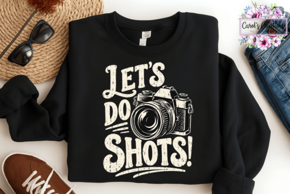 A black crewneck sweatshirt with a distressed white graphic that reads 'LET'S DO SHOTS!' alongside a camera illustration, styled in a flat lay with sunglasses, a straw hat, jeans, a shoe, and a floral 'Carol's Creations' logo in the corner.