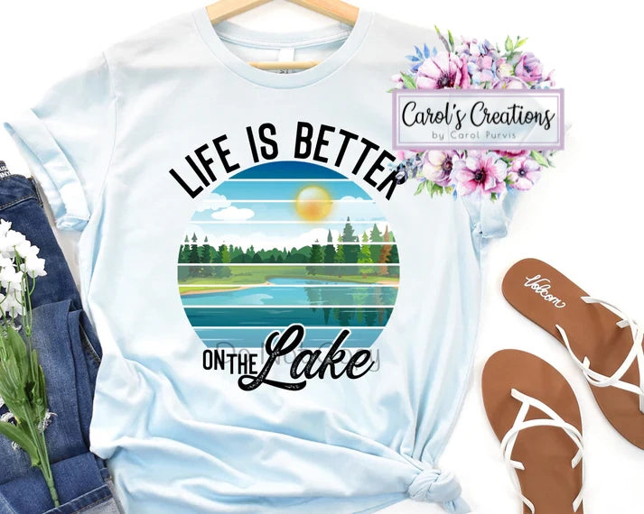 White t-shirt with 'Life is Better on the Lake' design, floral decorations, and 'Carol's Creations' branding.
