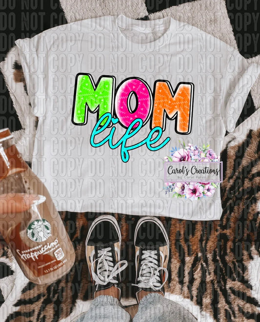 White t-shirt with 'Mom life' text, placed on a patterned surface with a bottle of coffee and shoes.