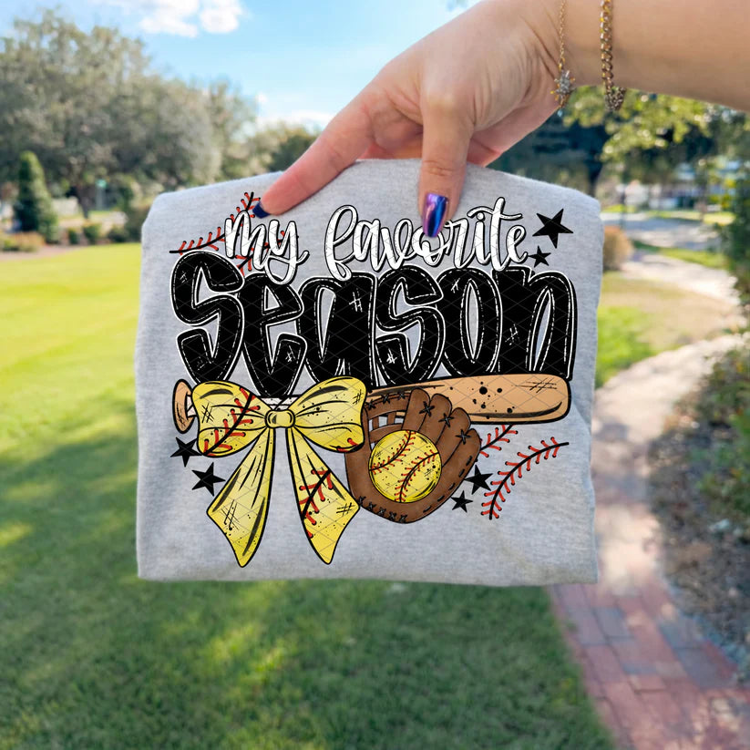 Gray sweatshirt with 'My Favorite Season' design held by a hand outdoors.