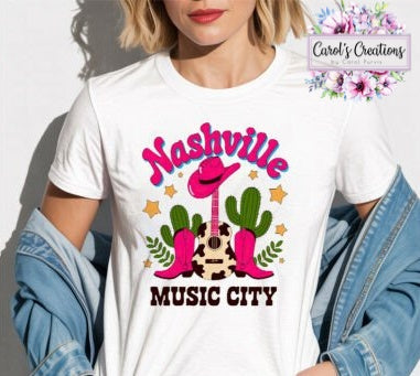 A woman wearing a white graphic t-shirt with a colorful 'Nashville Music City' western-themed design featuring pink boots, a guitar, and cacti. She has a denim jacket off her shoulders and is posed against a light gray wall.