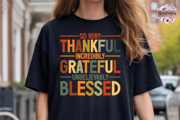 A woman wearing a black graphic t-shirt with a colorful text design that reads 'SO VERY THANKFUL INCREDIBLY GRATEFUL UNBELIEVABLY BLESSED'. The background is a blurred outdoor street scene.