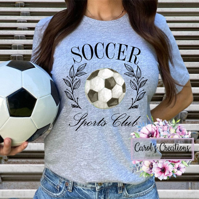 Person wearing a gray t-shirt with 'Soccer Sports Club' design, holding a soccer ball.
