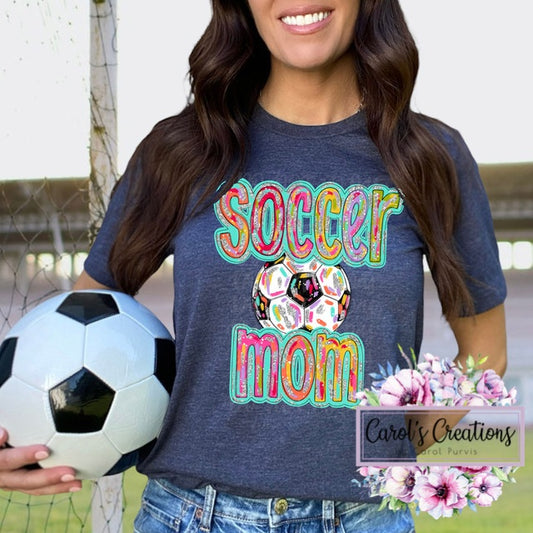Woman wearing a 'Soccer Mom' shirt holding a soccer ball on a sports field.