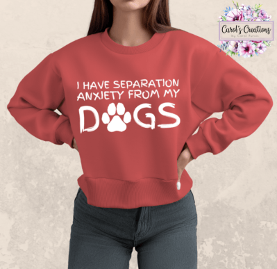 A woman wearing a coral red crewneck sweatshirt with a white graphic that reads 'I HAVE SEPARATION ANXIETY FROM MY DOGS' with a paw print graphic, paired with dark gray jeans. The background is a textured light beige wall, and a 'Carol's Creations' logo is visible in the top right corner.