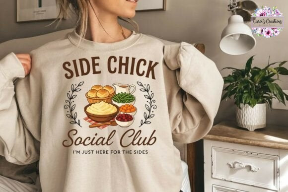 A woman wearing a beige crewneck sweatshirt with a graphic print that reads 'SIDE CHICK Social Club I'M JUST HERE FOR THE SIDES' and shows a bowl of dip, crackers, and a drink. The photo is taken in a cozy bedroom setting with a lamp and a plant visible in the background.