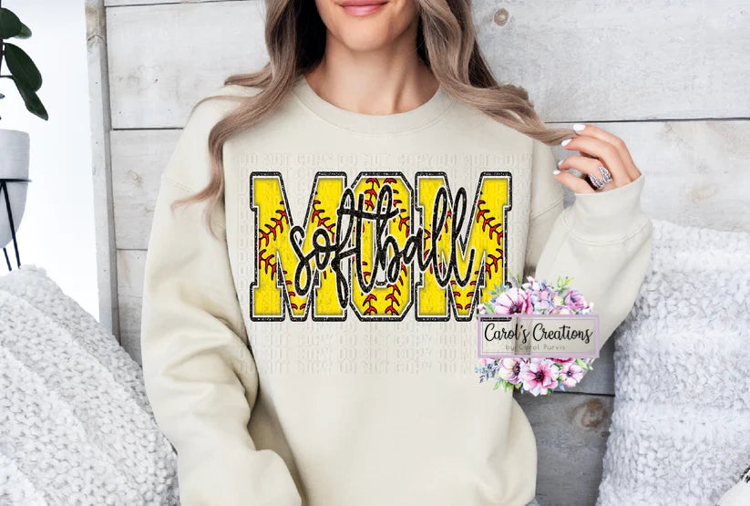 Woman wearing a cream sweatshirt with 'SOFTBALL MOM' text, sitting on a textured surface.