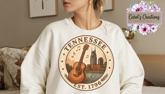 A woman wearing an oversized cream sweatshirt with a circular Tennessee state-themed graphic on the front, featuring an acoustic guitar, city skyline, and the text 'TENNESSEE EST. 1796'.