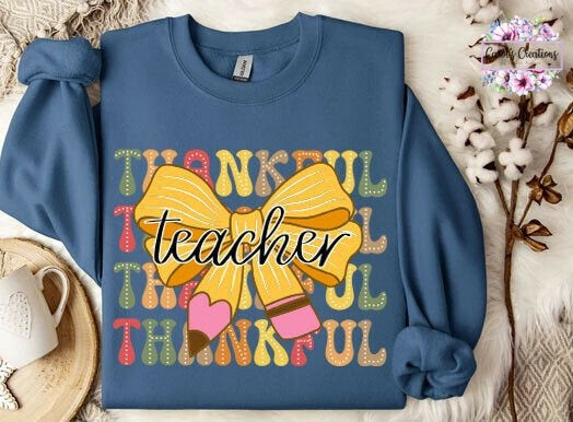 A folded long-sleeve crewneck sweatshirt in slate blue, featuring a colorful graphic with the word 'THANKFUL' in autumn-themed colors, a large yellow bow, and the word 'teacher' in black script. The sweatshirt is styled in a flat lay with a white mug, a woven coaster, cotton stems, and a white faux fur background.