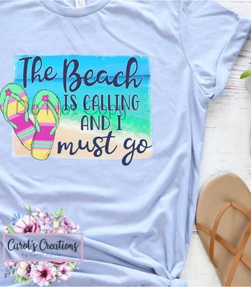 Light blue t-shirt with 'The Beach Is Calling And I Must Go' text and flip-flops design, placed on a wooden surface.