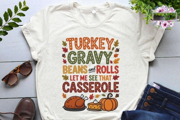 An off-white short-sleeve graphic t-shirt laid flat on a light gray wood plank surface. The shirt features a colorful autumn-themed graphic with the text 'TURKEY GRAVY BEANS and ROLLS LET ME SEE THAT CASSEROLE' in orange, green, and brown lettering, surrounded by leaves, with illustrations of a turkey, cheese, and a pumpkin at the bottom.